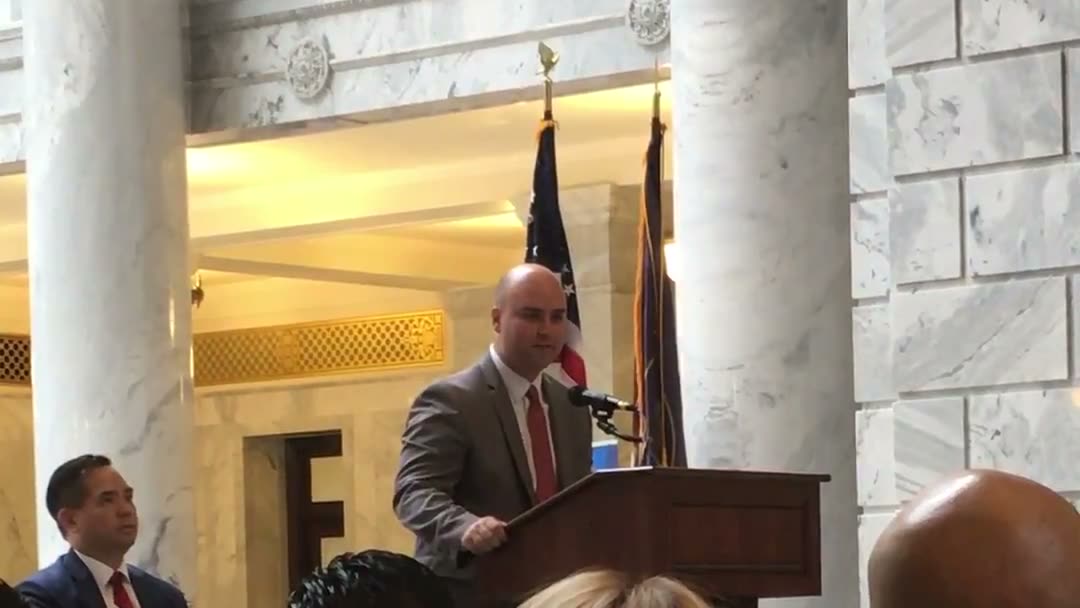 Daniel Souza at the podium in the Utah State Capitol rotunda, U.S. and Utah flags behind him, Attorney General Sean Reyes seated to the left